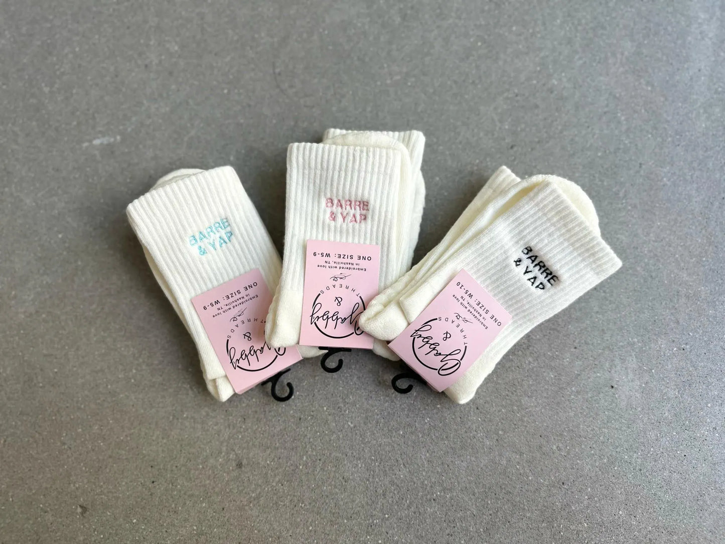 three pairs of white grip socks are laying on a concrete floor. All three pairs of socks have the words "Barre & Yap" embroidered on them in different thread colors (pink, light blue and black).