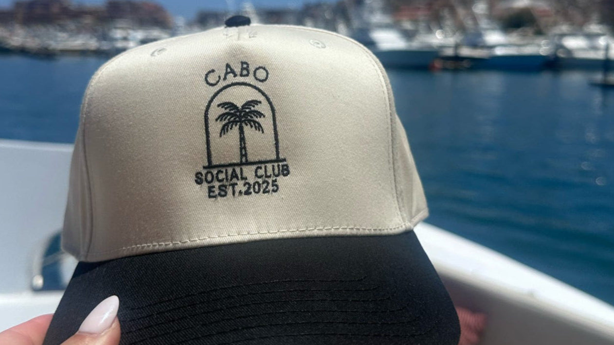 Beige and black cap with 'Cabo Social Club' logo held by a hand with a scenic background of water and mountains.
