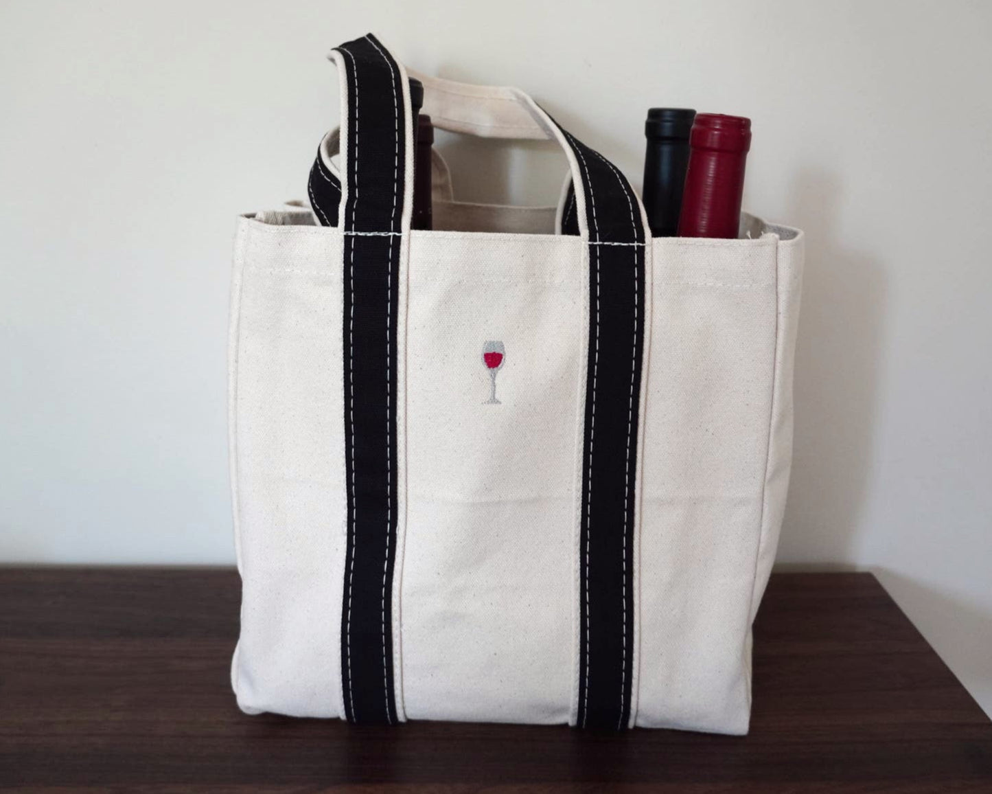 White tote bag with black handles and a small red wine glass design, containing wine bottles, on a wooden surface