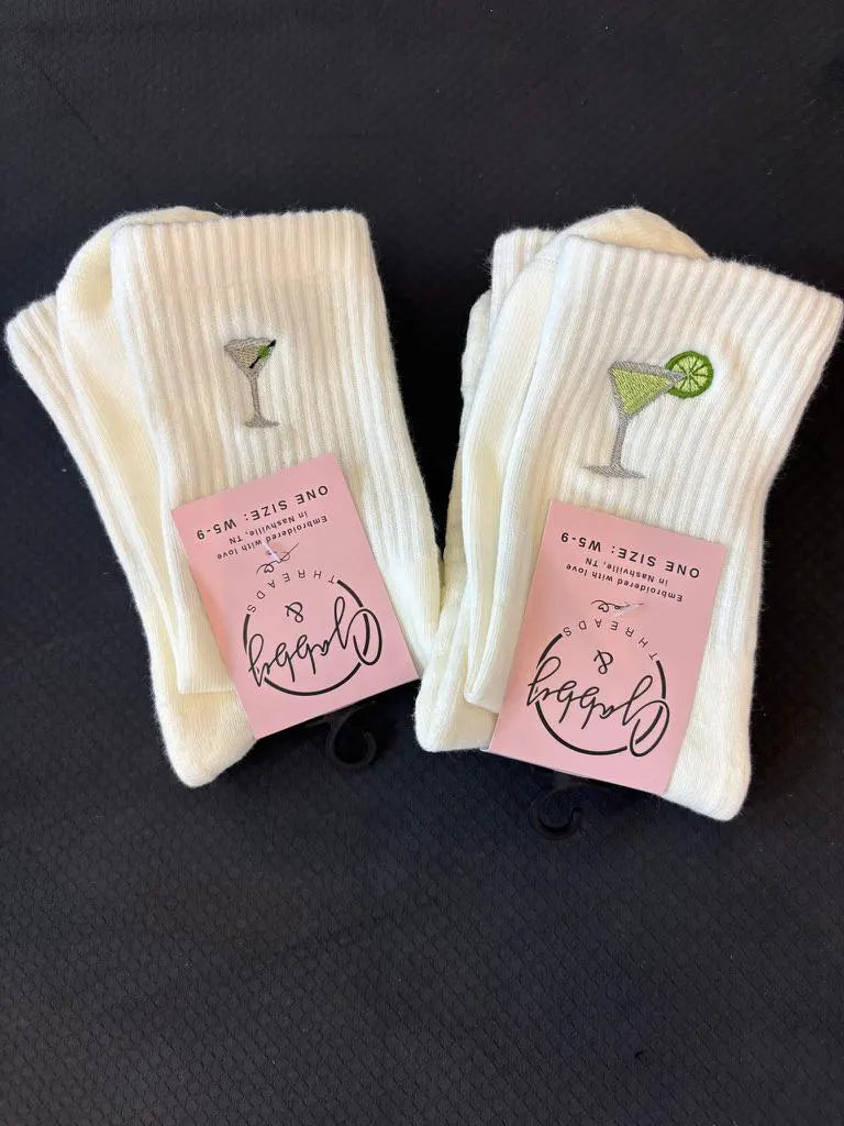 two pairs of white grip socks laying on a black yoga mat. One pair of socks has a dirty martini embroidered on it and the other one has a margarita
