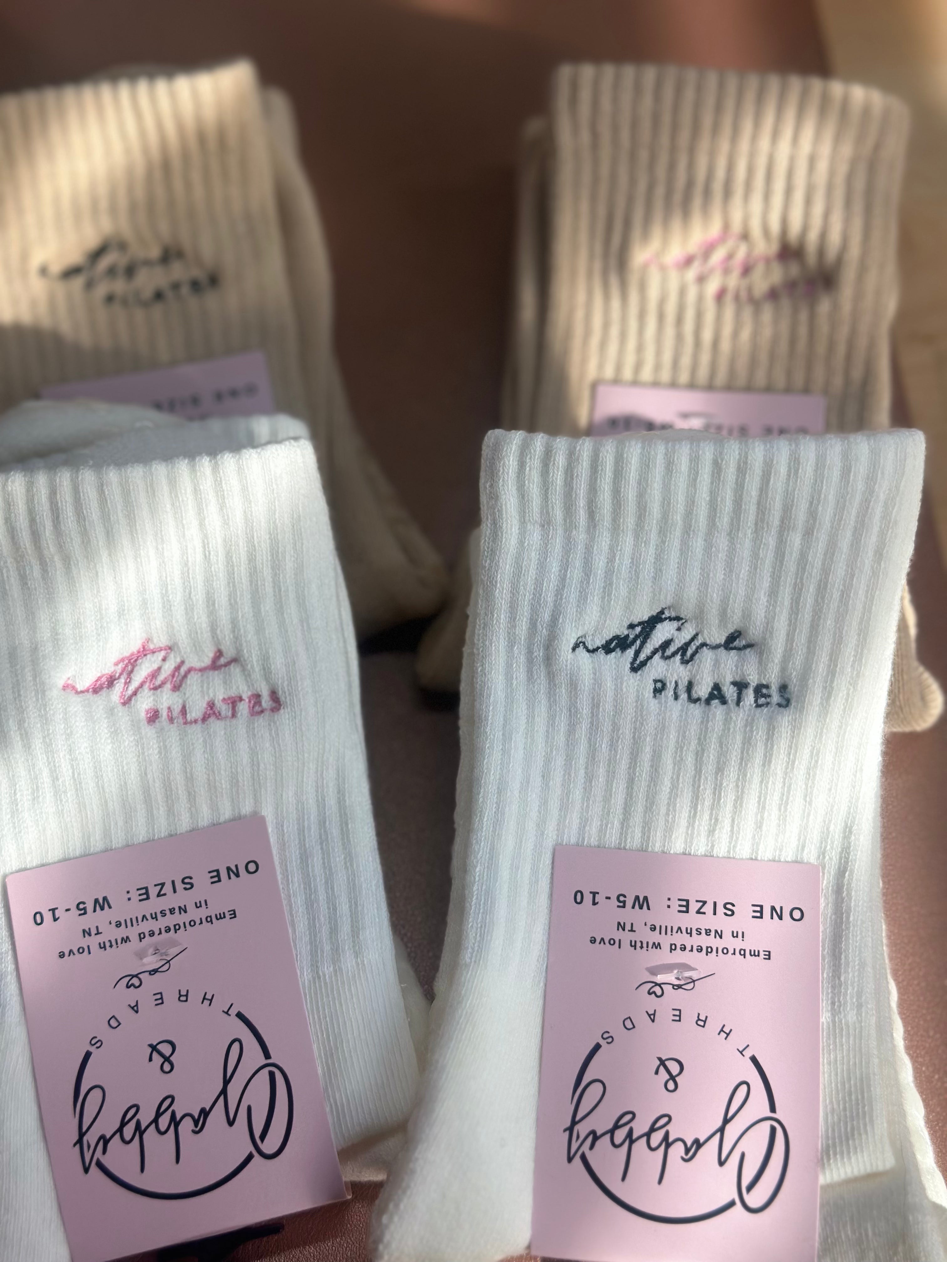 White and tan socks with 'Native PILATES' branding and pink packaging in the foreground.