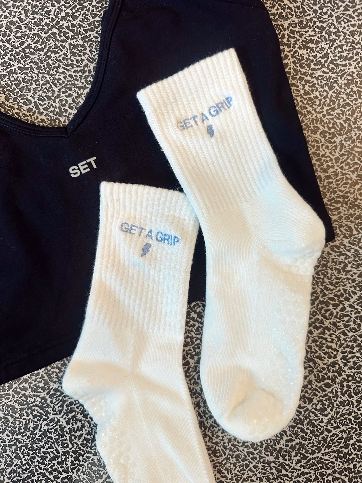 White grip socks socks with 'GET A GRIP' text embroidered on them, The socks are laying on on a black SET Active tank on a patterned surface.