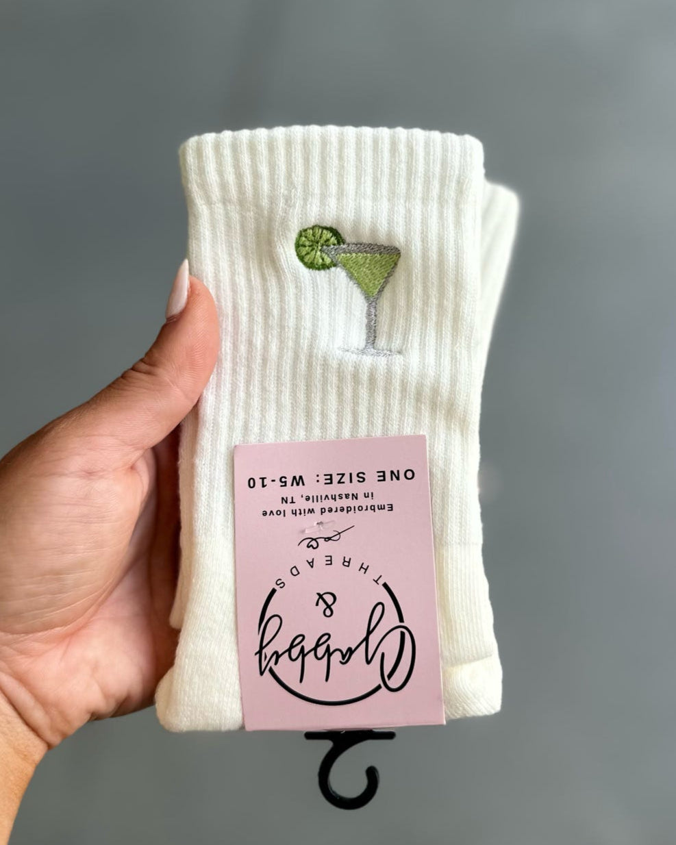 Hand holding a pack of white socks with an embroidered green margarita with a pink label against a gray background