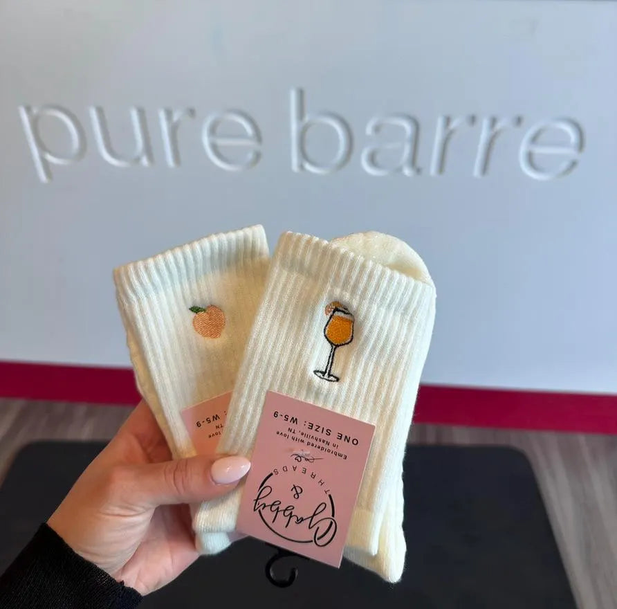 two pairs of white socks at Pure Barre. One sock has a peach embroidered on it and the other pair of socks has an aperol spritz embroidered on it