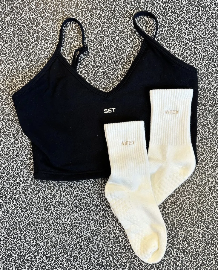 white grip socks with the words "wifey" embroidered on them in beige thread. The socks are laying on a yoga mat on top of a black workout top from the brand Set Active.