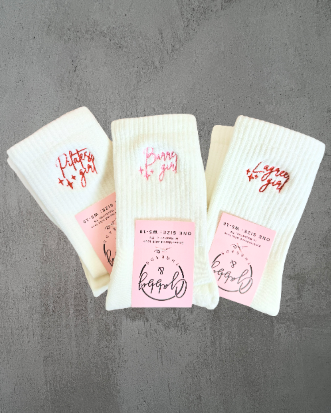 Three pairs of white grip socks with the words "pilates girl" "lagree girl" and "barre girl" embroidered on them