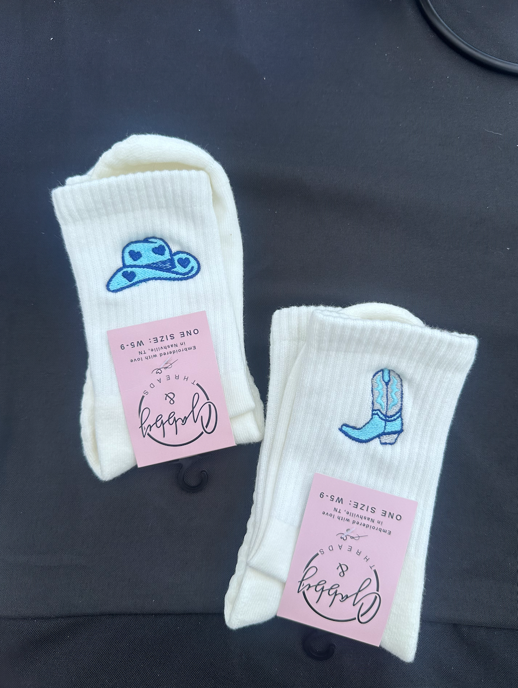Two pairs of white socks with blue cowgirl hat and cowboy boot designs and pink labels on a dark background