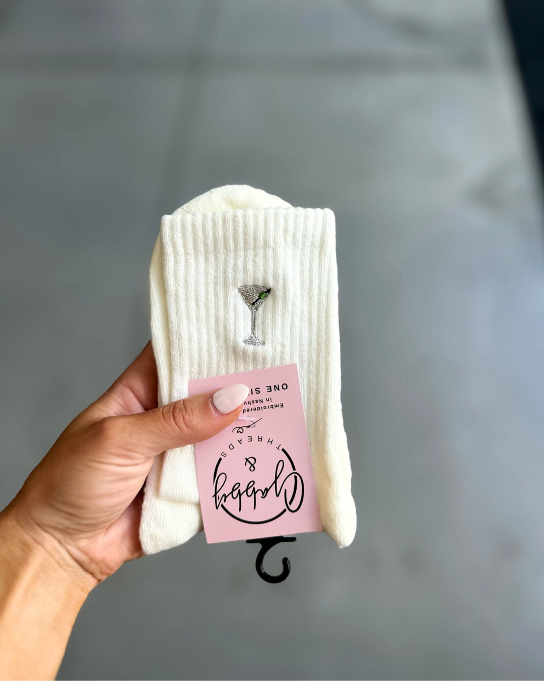 White socks with an embroidered dirty martini design held by a hand against a gray background