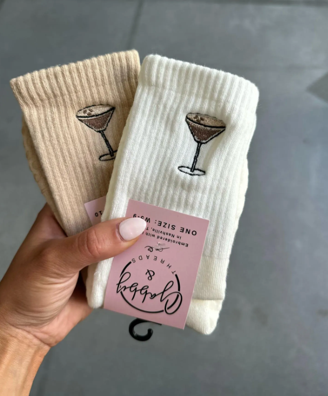 White and beige grip socks with espresso martini embroidered on them. The socks are held up by a hand against a grey tile floor.