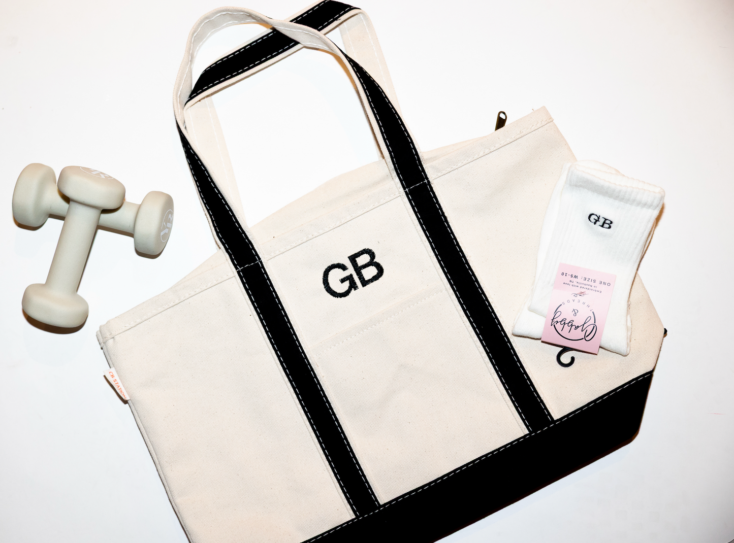 White tote bag with black straps and embroidered initials dumbbell, and product package on a white background with