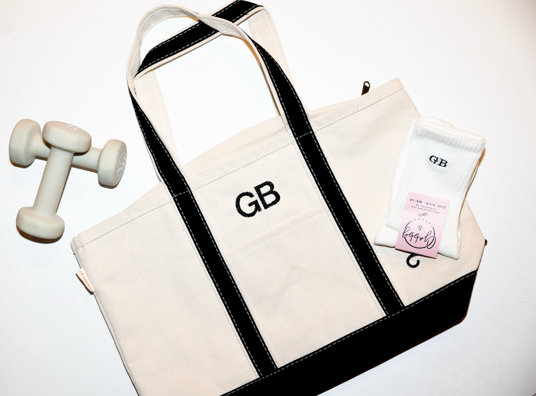 White tote bag with black straps and embroidered initials dumbbell, and product package on a white background with