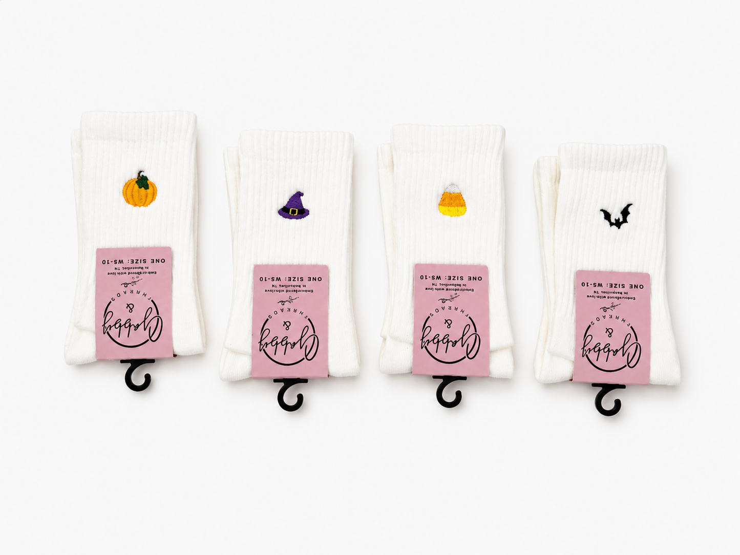 Four pairs of white ribbed socks displayed side-by-side on black metal hangers against a white background. Each pair features a small embroidered  Halloween seasonal graphic: a pumpkin, a witch hat, a candy corn, and a bat, respectively.