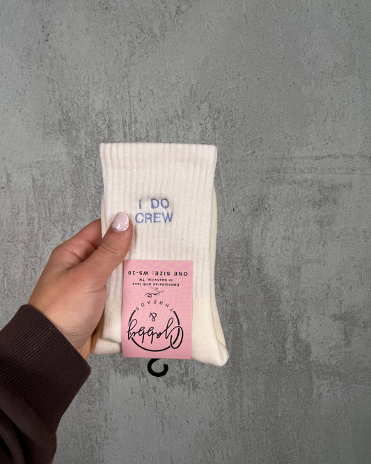 a hand holds up a pair of white grip socks with the words "I do Crew" embroidered on it against a grey background