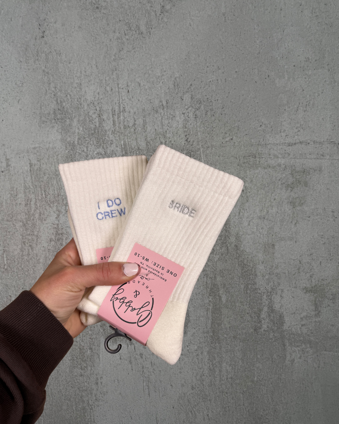 a hand holds up two pairs of socks, one with the word "bride" and the other with the word "I do crew" against a gray background