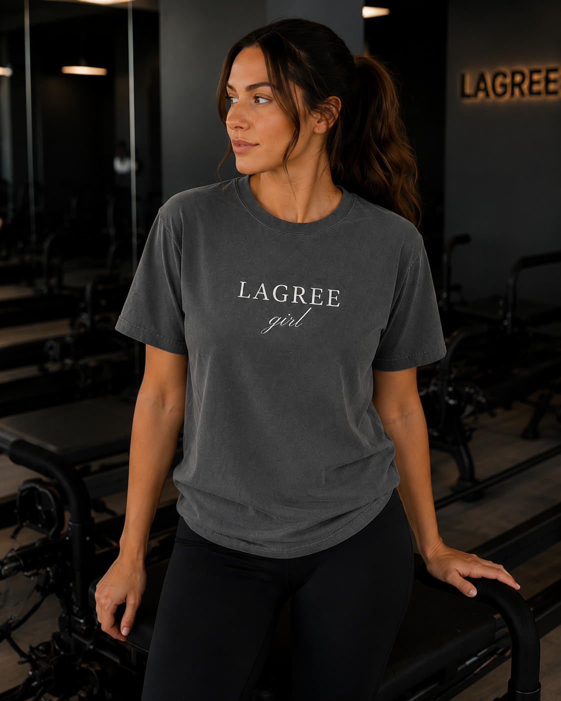 Woman wearing a gray t-shirt with 'LAGREE girl' text in a gym setting