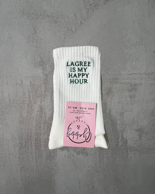 A pair of white embroidered socks that say "LAGREE IS MY HAPPY HOUR" embroidered in green. 