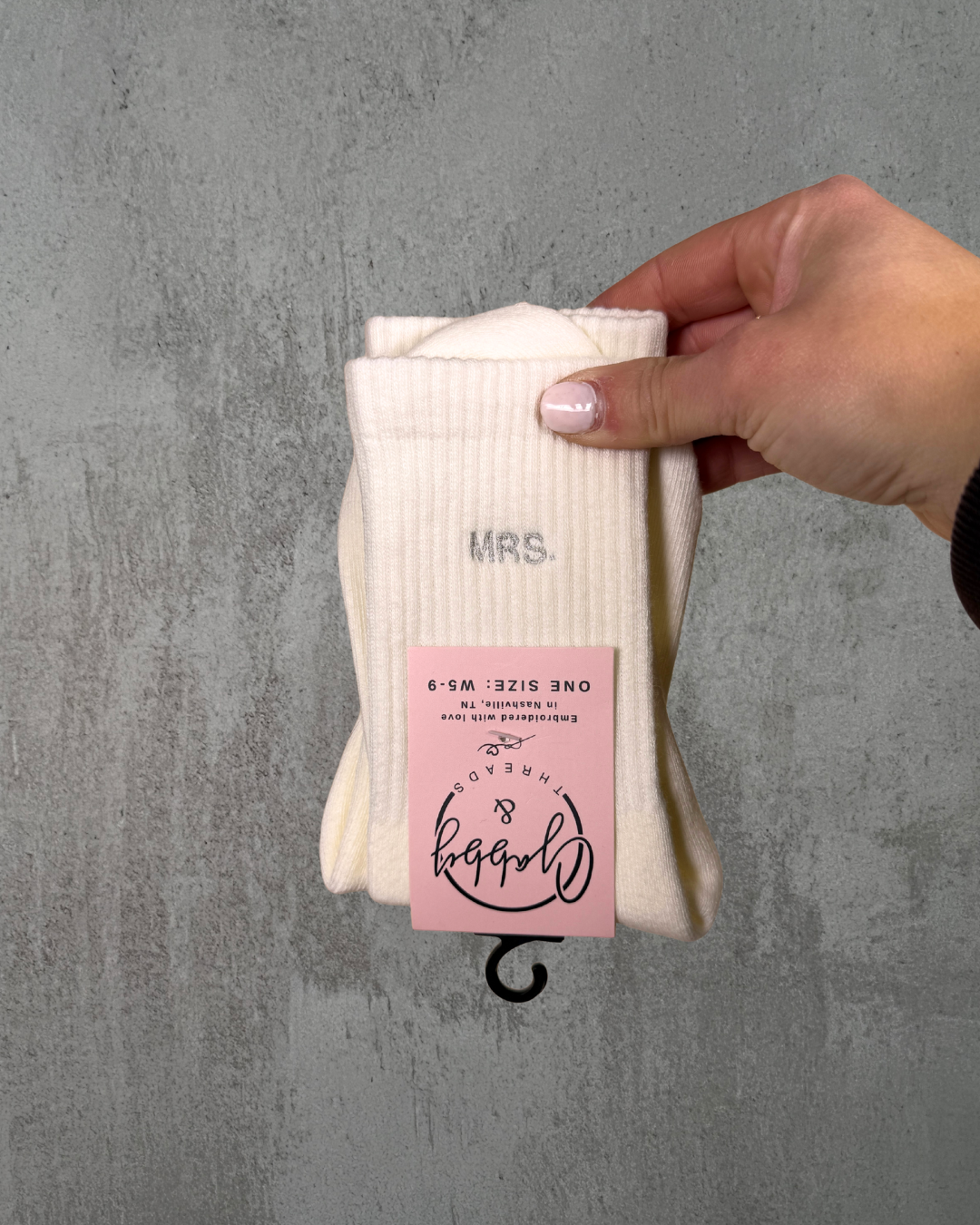 A Hand is holding a pair of socks with the text "Mrs." embroidered on it against a gray background