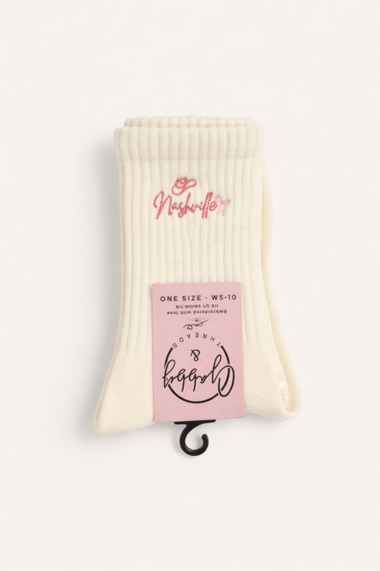 a pair of white grip socks for pilates with the word "nashville" embroidered on them with a pink cowboy hat and bow