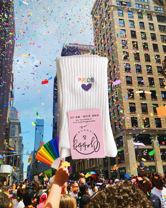 a pair of white socks with the word "pride" embroidered on them in rainbow colors against a background with a pride festival