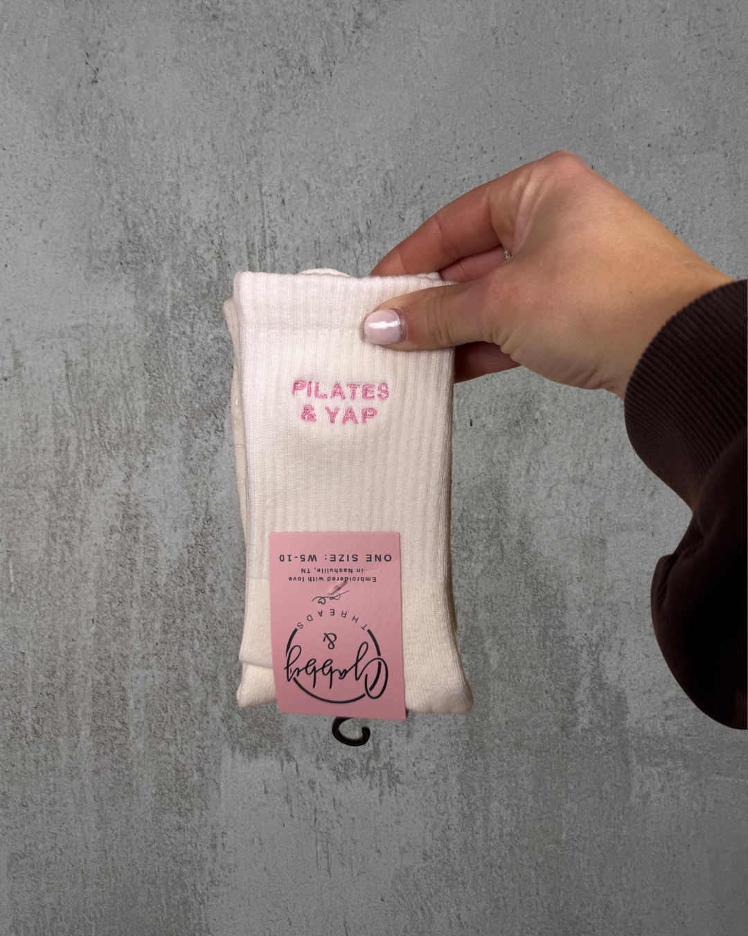 a hand holds up a pair of white grip socks with the text "pilates & Yap" embroidered on it against a grey background