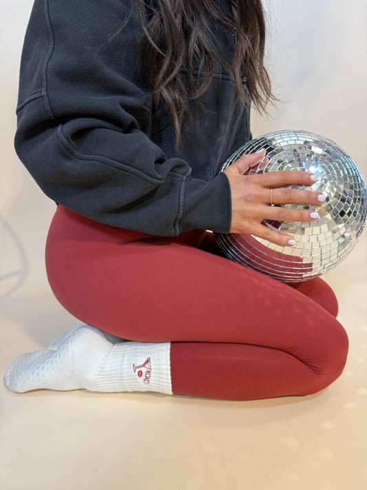A girl is wearing white grip socks for pilates on her feet that are themed for Valentine's day. She has a disco ball in her hands.