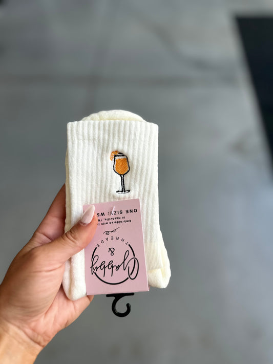 White socks with aperol spritz and pink tag held by a hand on a gray background