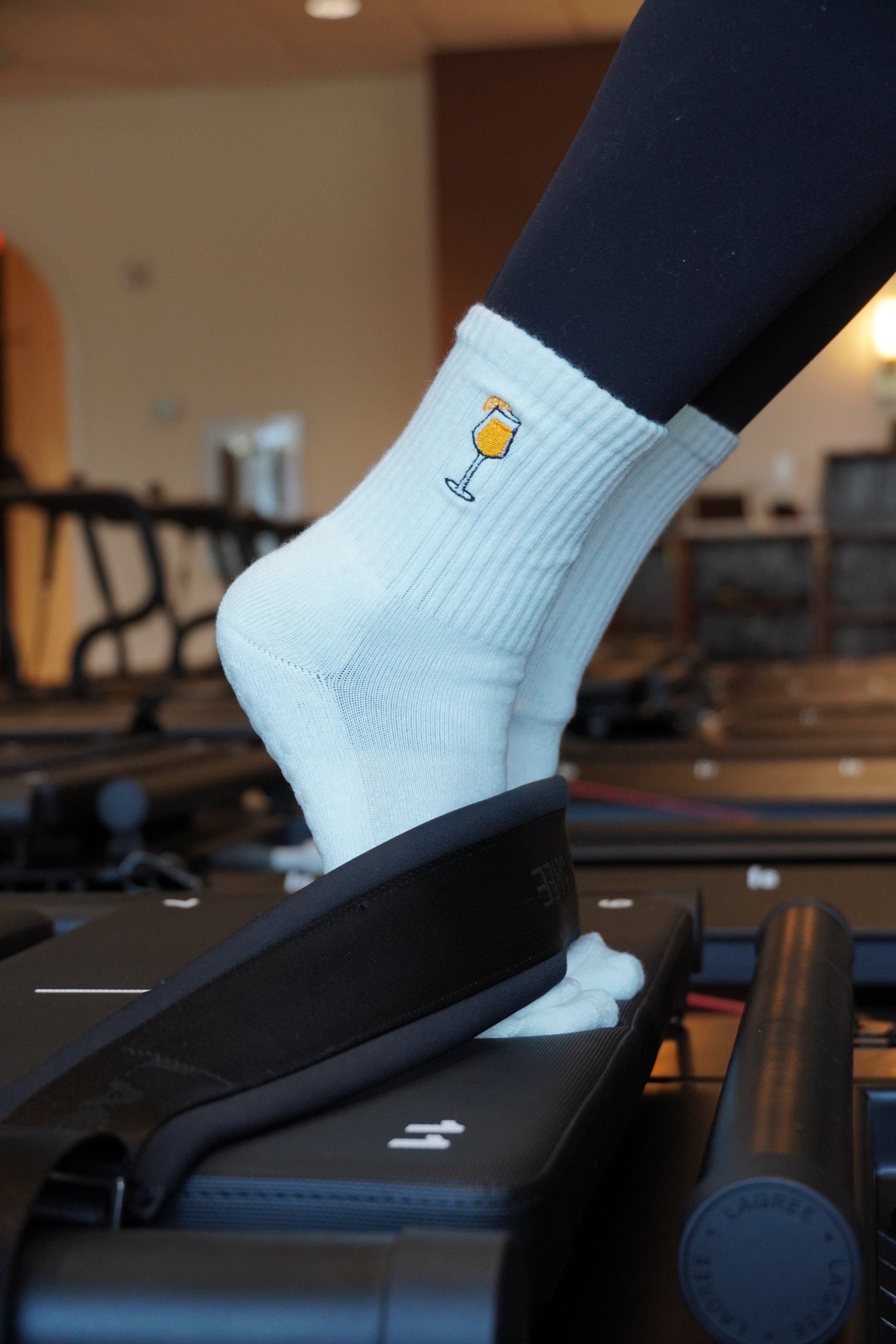 a person is wearing a pair of white grip socks with an embroidered Aperol Spritz design on a pilates reformer