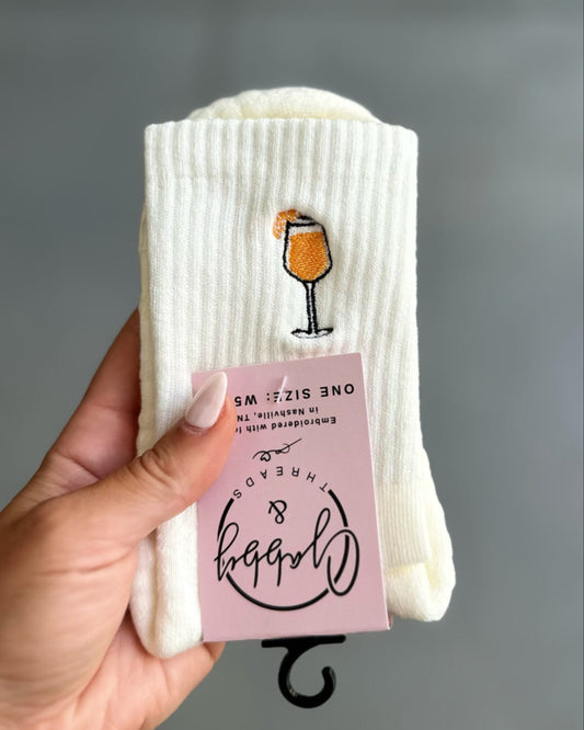 White socks with aperol spritz and pink tag held by a hand on a gray background
