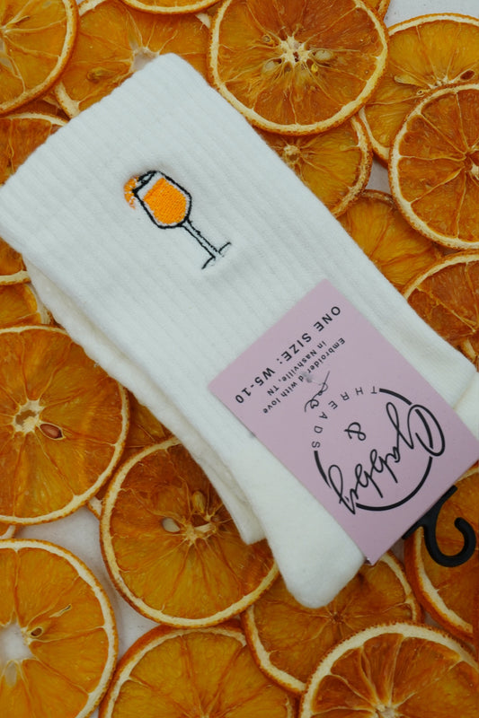 a pair of aperol spritz embroidered socks laying across a bunch of orange slices