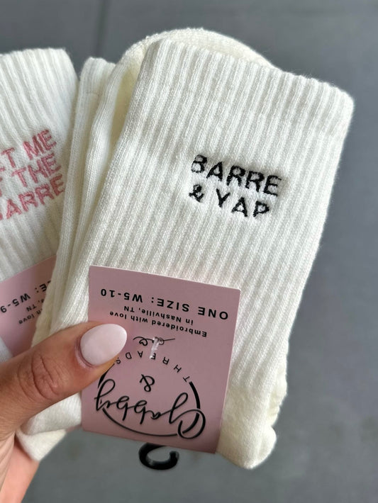 White socks with 'Barre & Yap' text held by a hand against a gray background