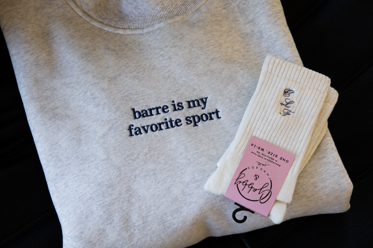 a folded mockneck sweatshirt featuringt the words "barre is my favorite sport" with a pair of grip socks laying on top 