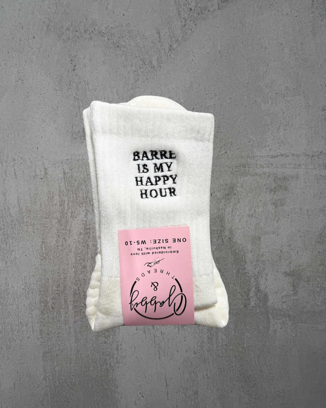 A pair of white embroidered socks that say "BARRE IS MY HAPPY HOUR" embroidered in black.