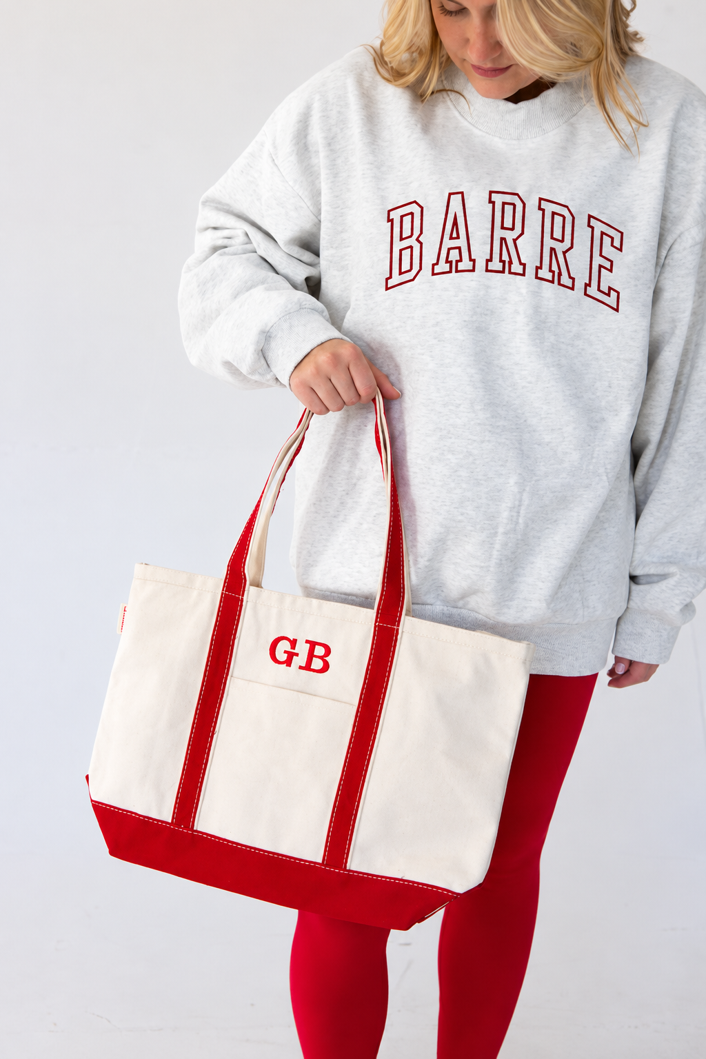a women is wearing a heather gray mockneck with the word "BARRE" embroidered on it in varsity font. Carrying a canvas tote bag with her initials embroidered on it
