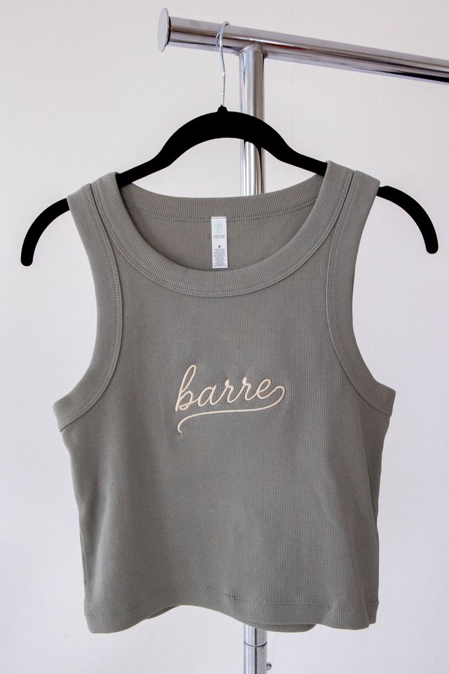 A sleeveless cropped tank top in a muted grayish green, hanging on a black hanger from a metal clothing rack. The front of the tank top features the word 'barre' embroidered in a light beige cursive script.