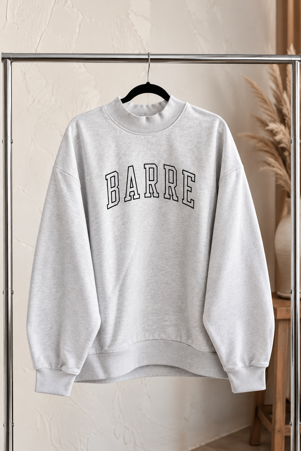 a heather gray mock neck sweatshirt is hanging on a clothing rack. The sweatshirt is embroidered with the word "barre" in varsity letters