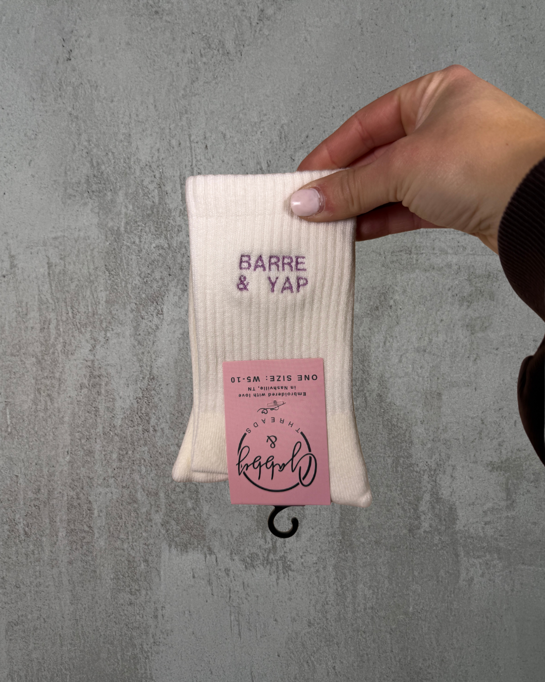 a hand holds up a pair of white grip socks with the text "barre & yap" embroidered on it against a grey background