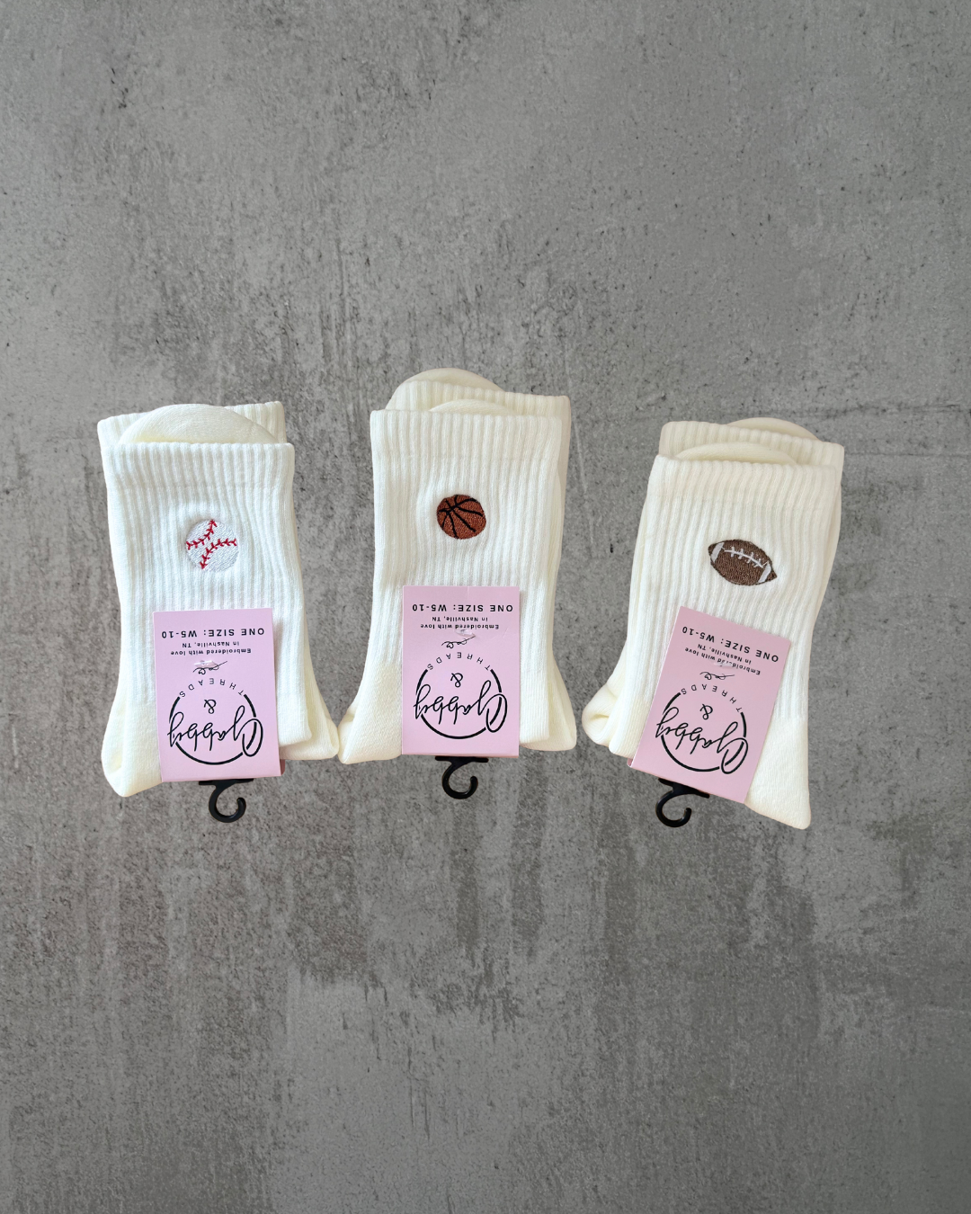 Three pairs of white socks, each with a different sports ball embroidered on them: a baseball, a basketball, and a football.  The socks are displayed on a gray background.