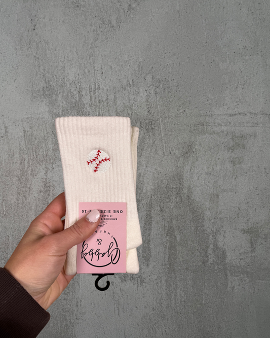 Hand holding a pair of white socks with an embroidered baseball on them against a gray background