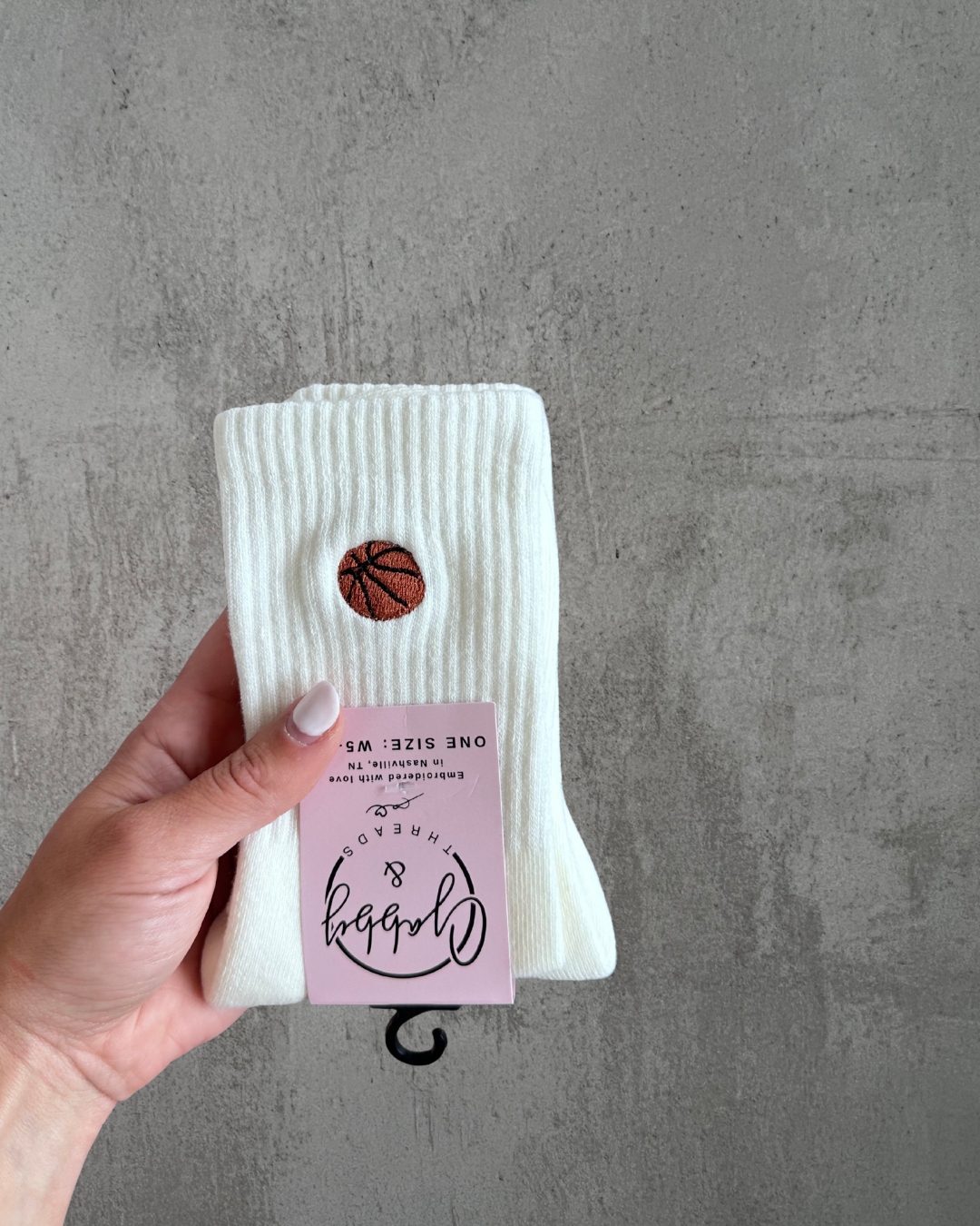 Off-white socks with a brown basketball embroidered on the front. They are held up by a hand against a grey background