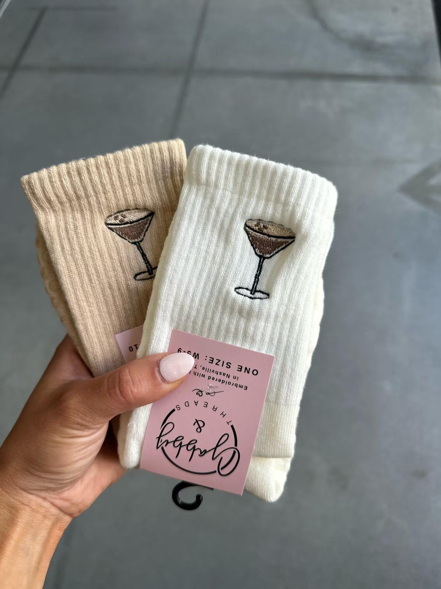White and beige grip socks with espresso martini embroidered on them. The socks are held up by a hand against a grey tile floor.