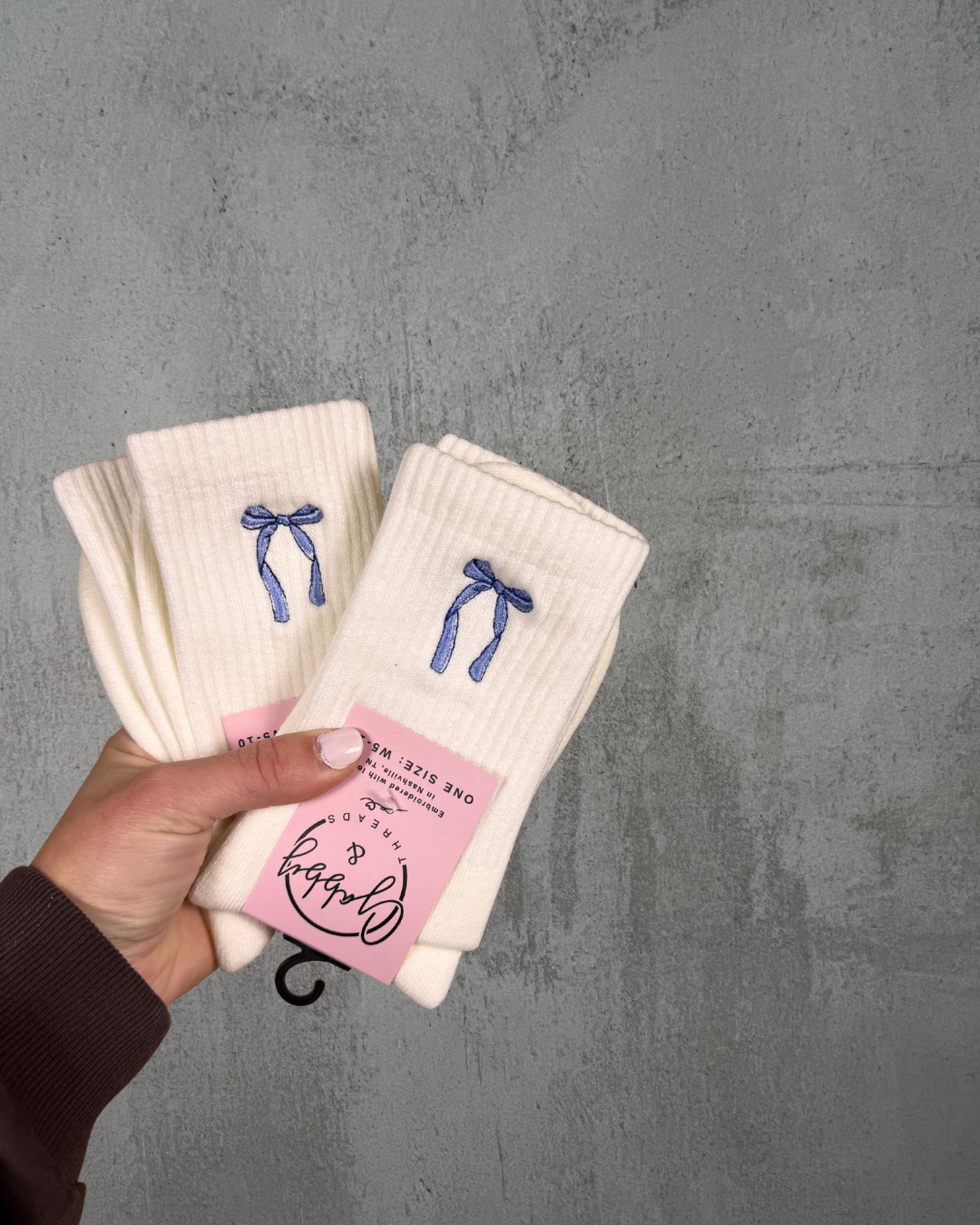 Two pairs of socks with embroidered bows held by a hand against a gray background