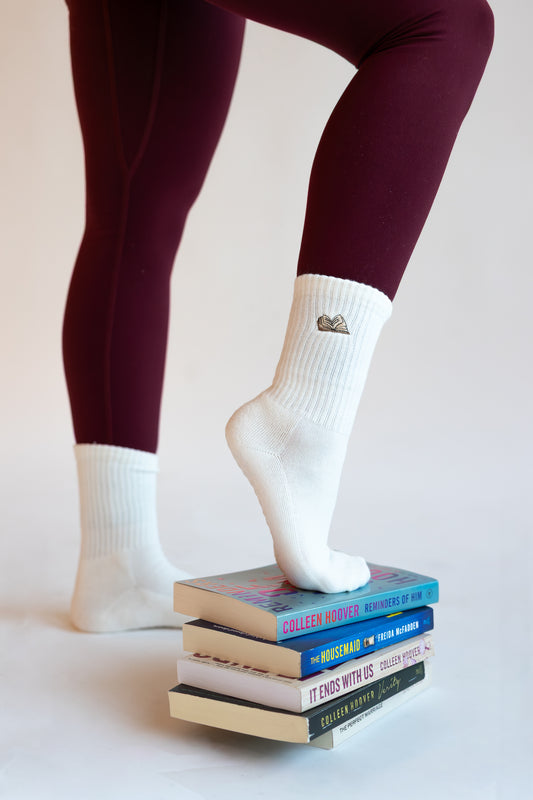 a person is wearing a pair of white crew socks with an embroidered book and has her foot propped up on a stack of books