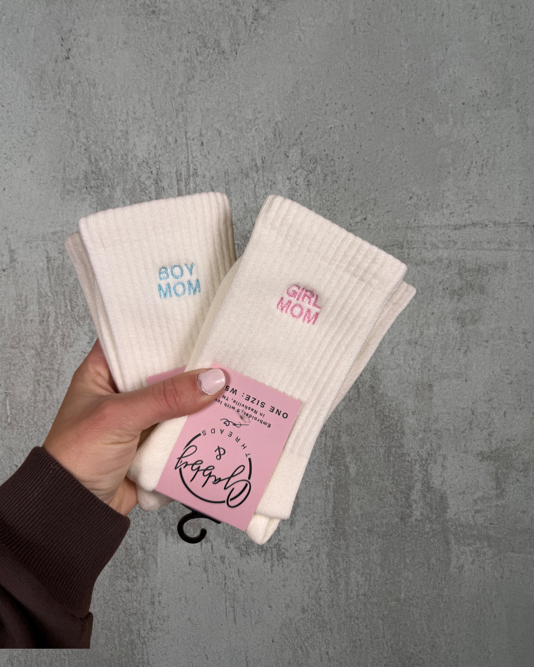 two pairs of white grip socks against a grey background. One pair of socks says "Boy mom" in blue thread and the other one says "Girl Mom" in pink thread color.