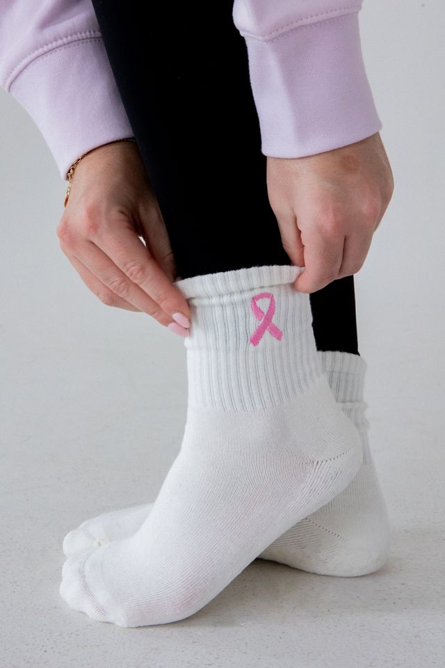 White ribbed ankle socks with a pink awareness ribbon on the cuff, shown being worn and adjusted on a person's foot against a light gray background.
