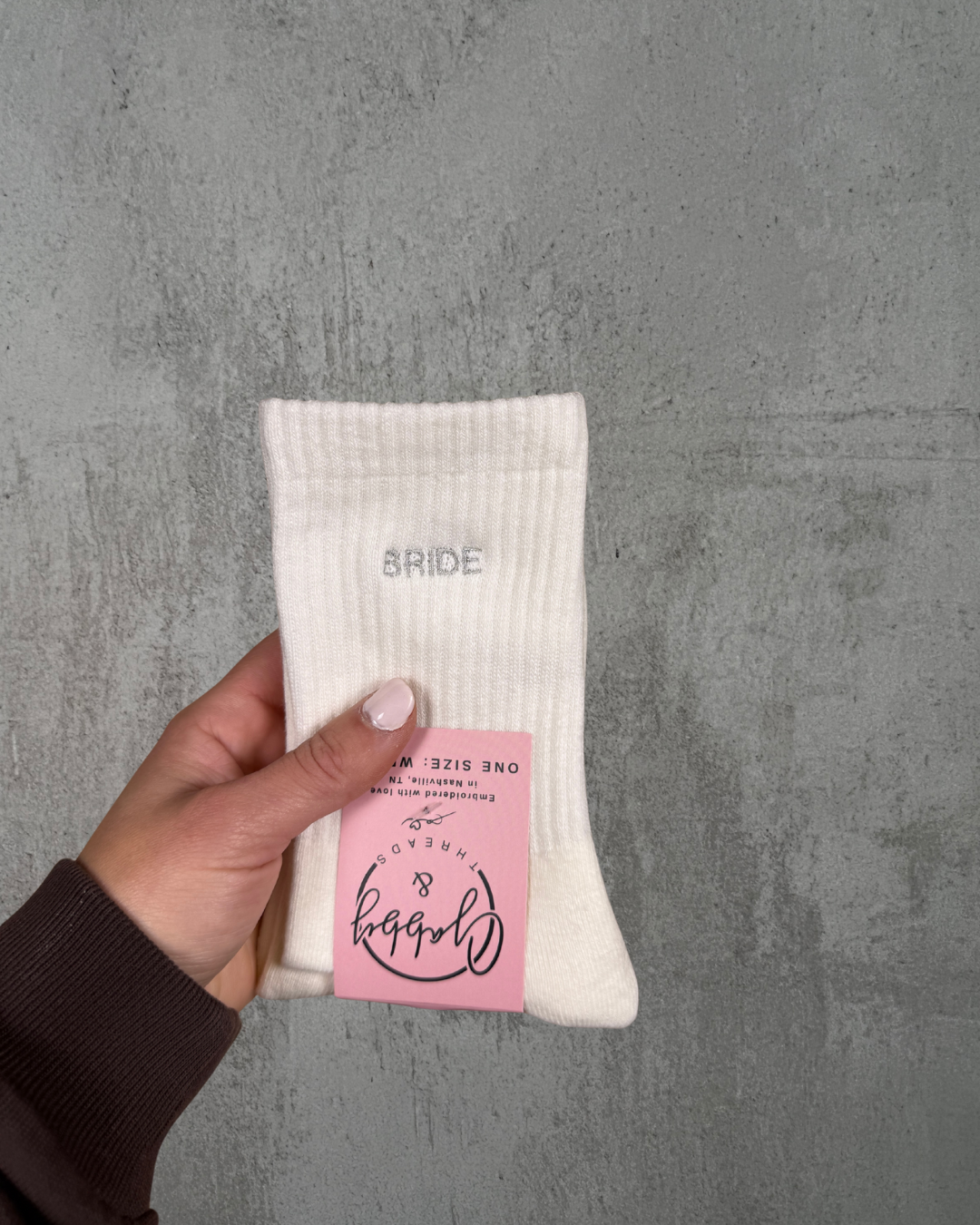 a hand holds up a pair of white grip socks with the word "bride" embroidered on it against a grey background
