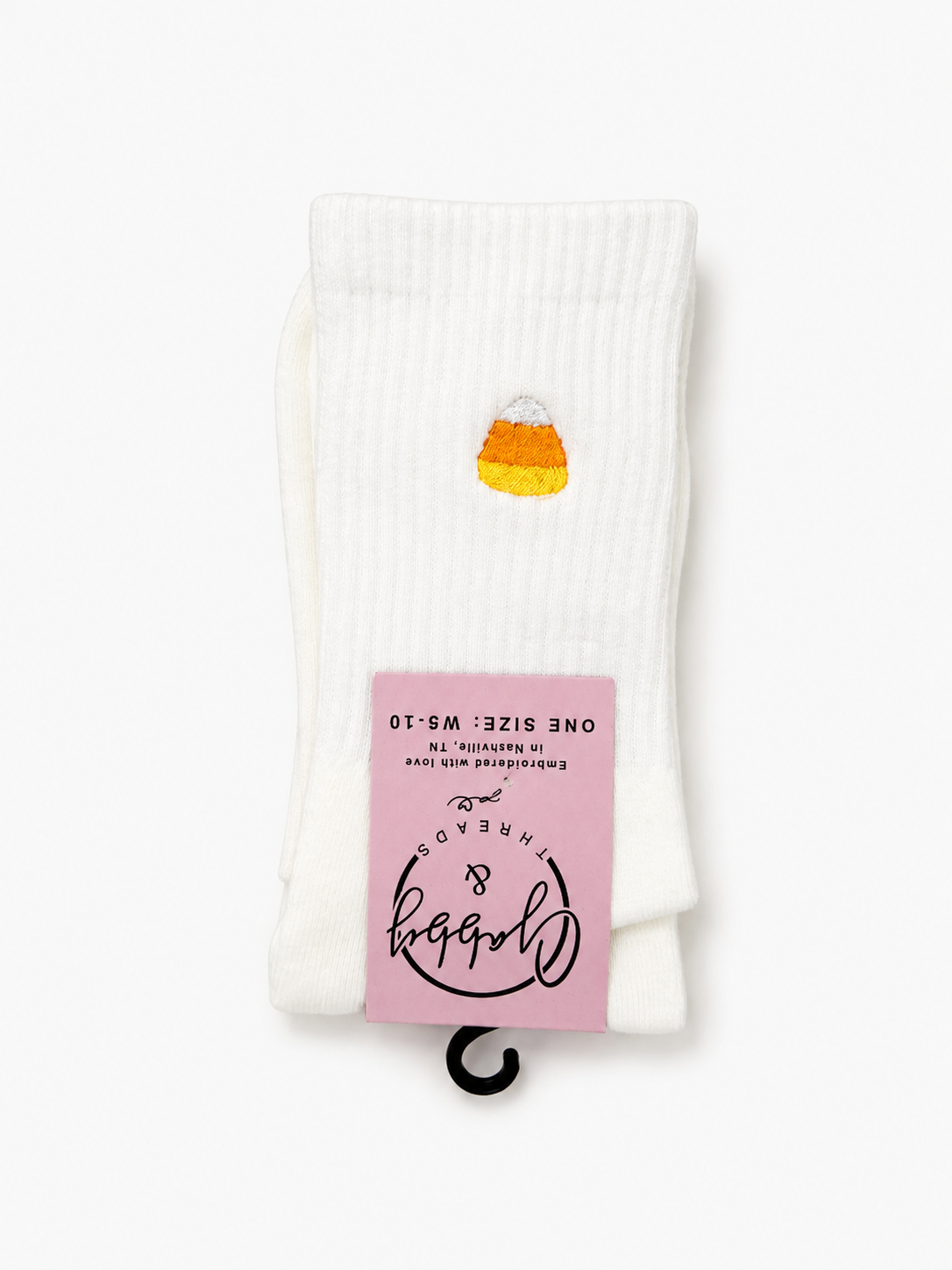 A pair of white ribbed crew socks with a small yellow and orange candy corn graphic on one sock, featuring a pink paper tag