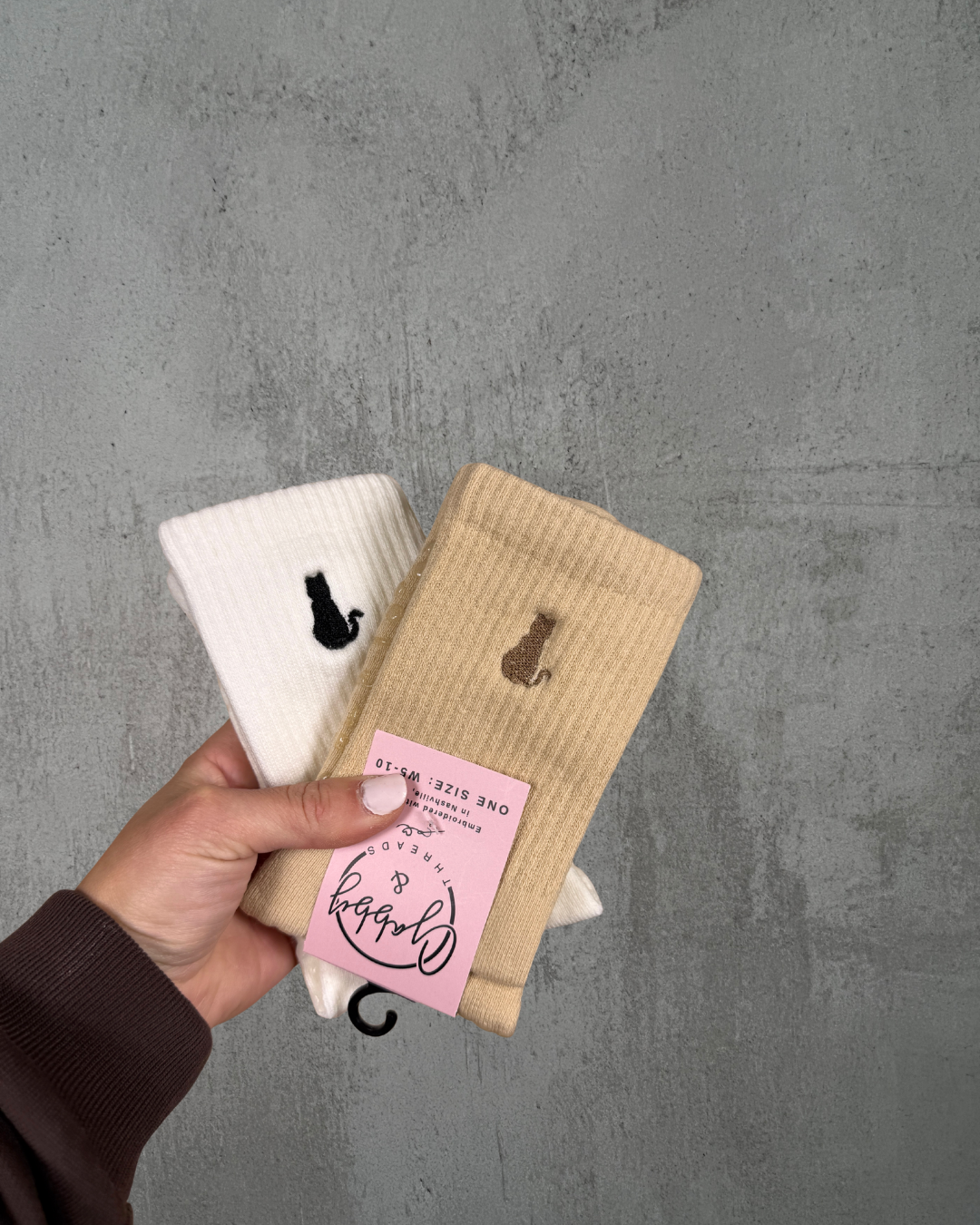 a hand holds up two pairs of socks, both socks have a cat embroidered on them against a gray background