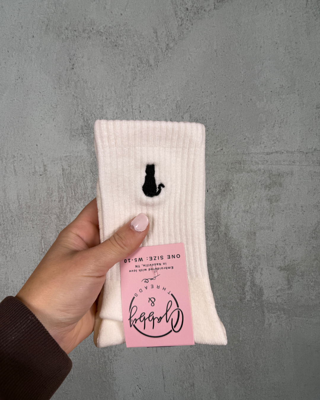 a hand holds up a pair of white grip socks with a cat embroidered on it against a grey background