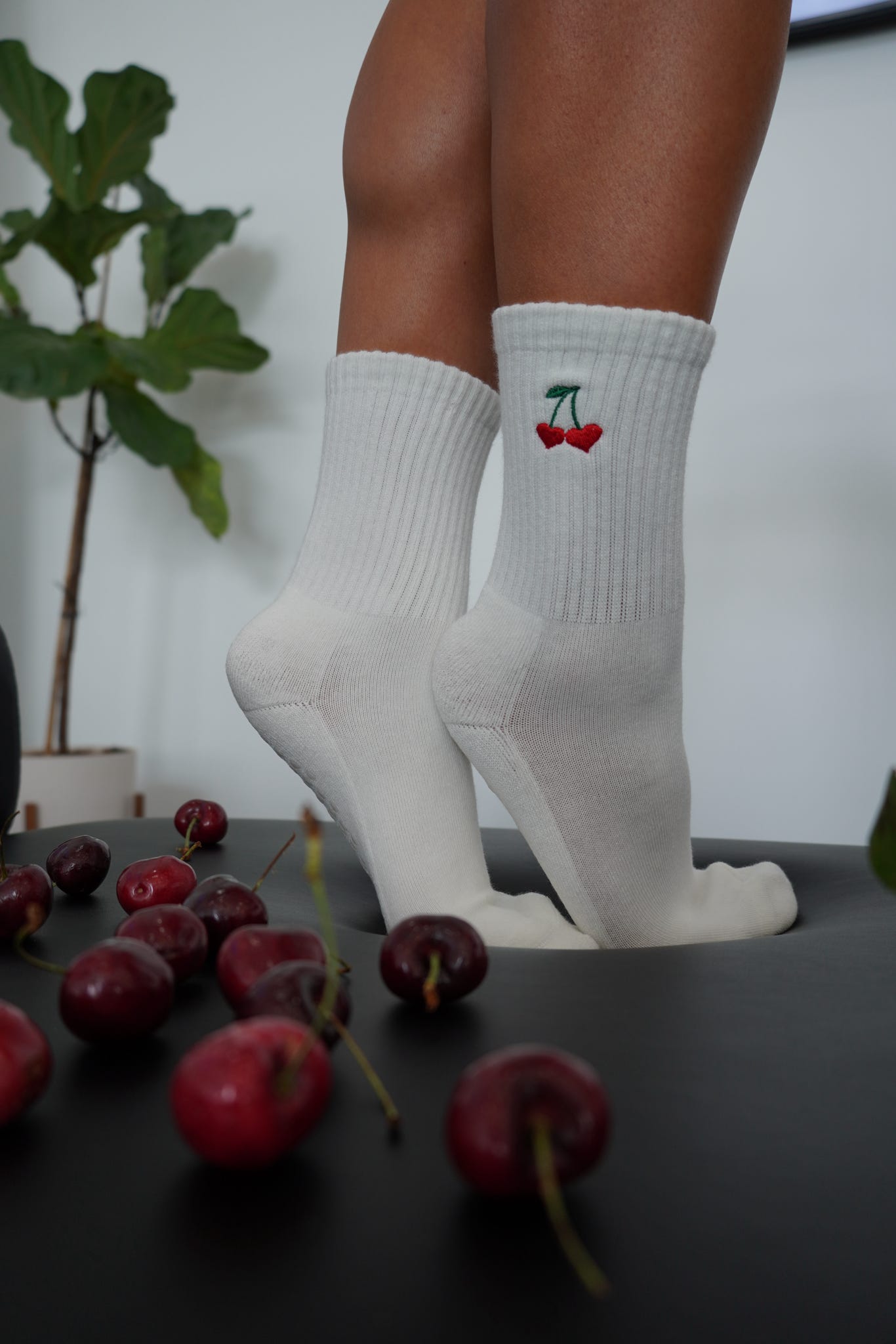 White socks with cherry design worn by a person, surrounded by cherries on a dark surface.