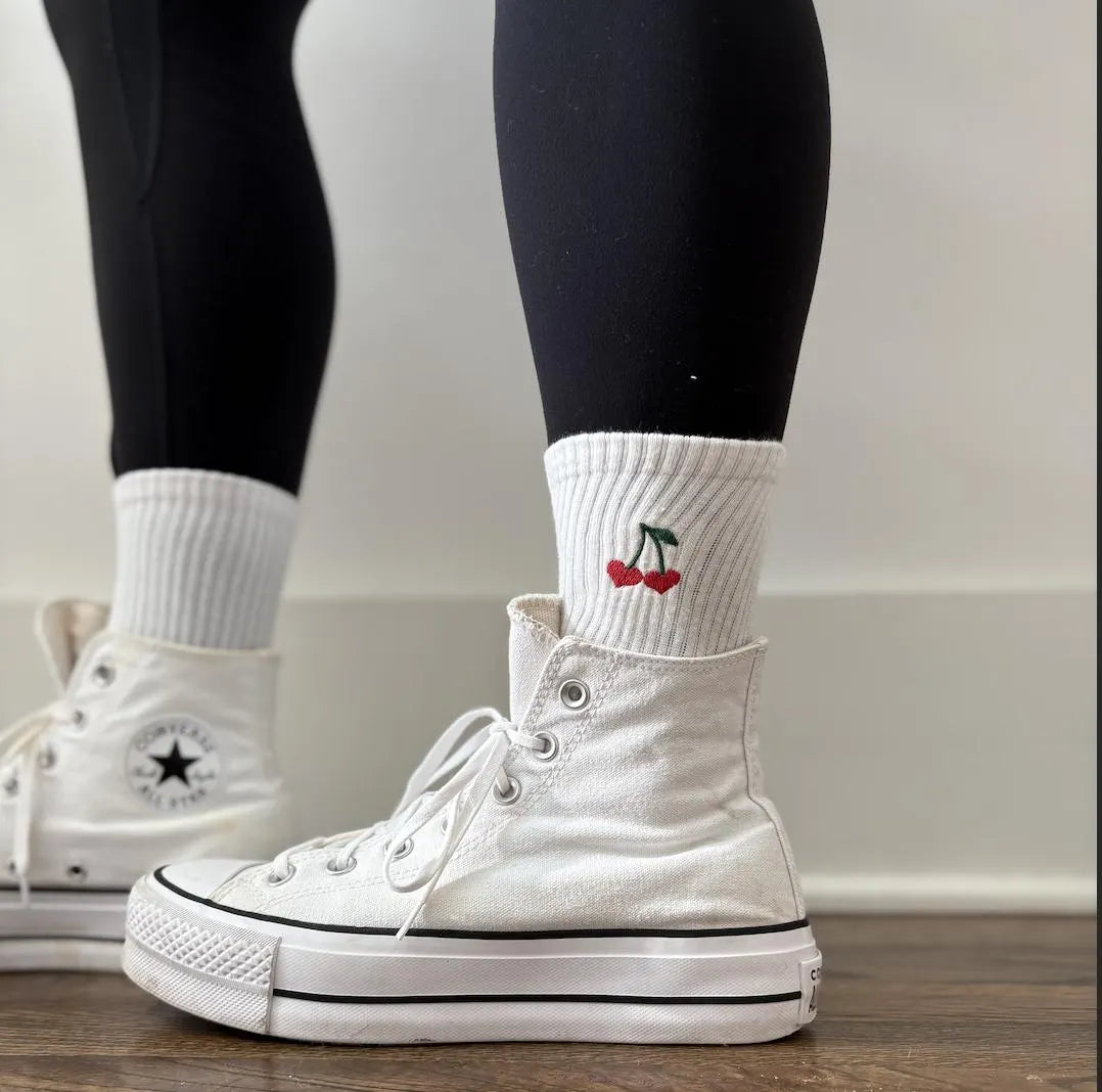 A person is wearing a pair of white high top converse with crew socks that have a pair of cherries embroidered on them
